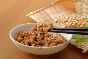 箸で持ち上げた糸引き納豆と大豆。白い器に盛られた納豆の奥に、ざるに入った大豆が置かれた和風イメージ画像